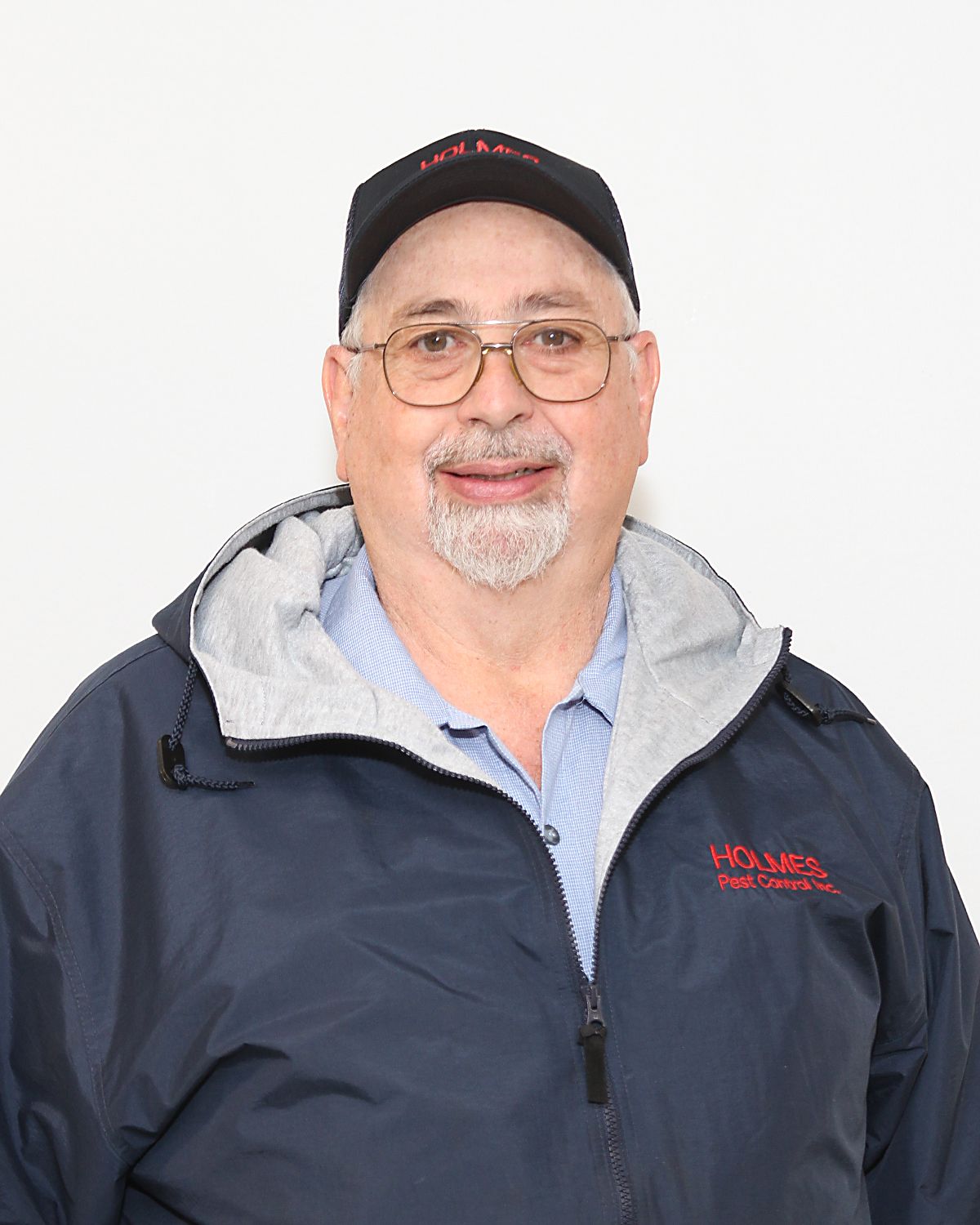 Man wearing glasses and a cap, zipped blue jacket, smiles at the camera.