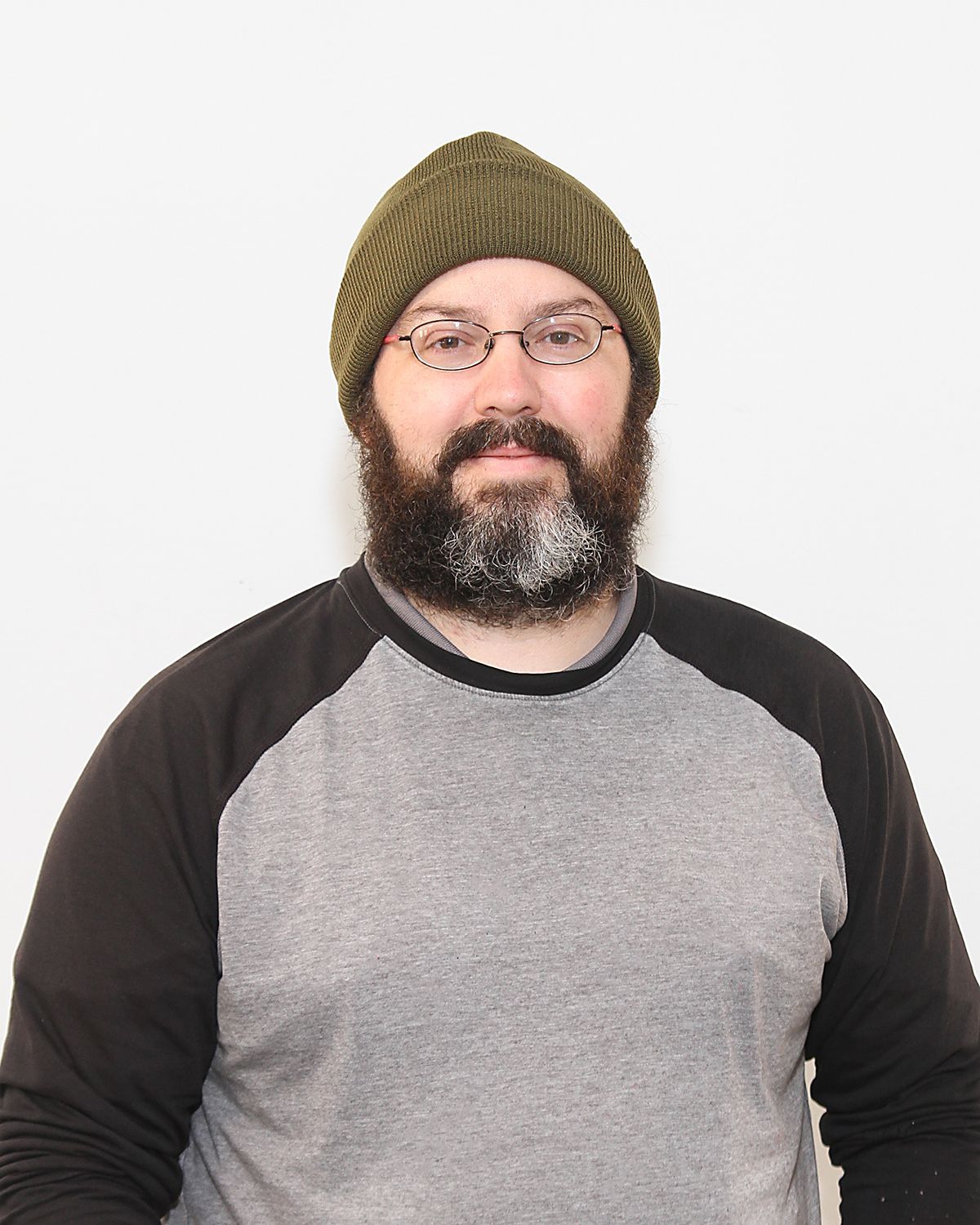Man with a full beard, wearing glasses and a green beanie, gray and black raglan shirt, standing against a white background.