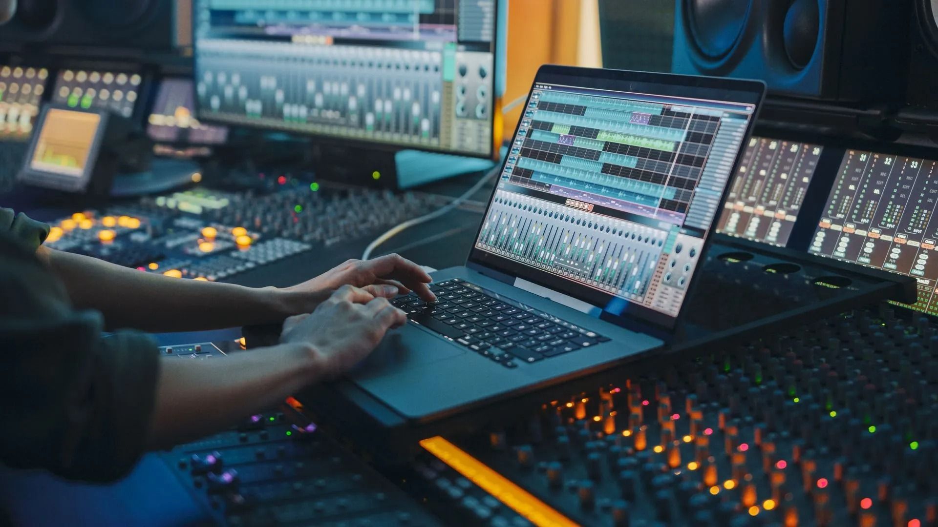 Hands using a laptop in a music production studio with mixing consoles and glowing audio controls.