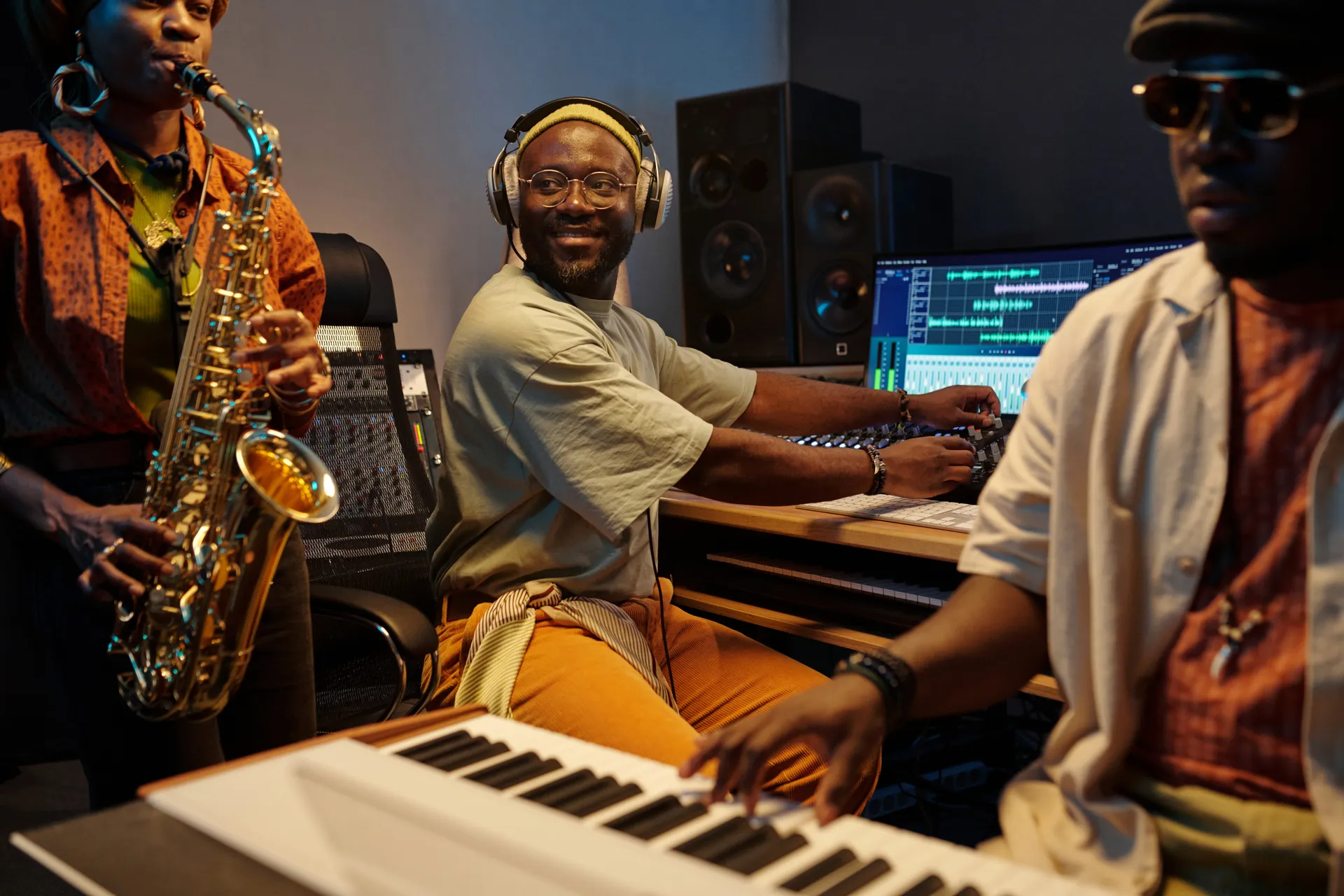Three musicians in a studio, with saxophone, keyboard, and mixing equipment.