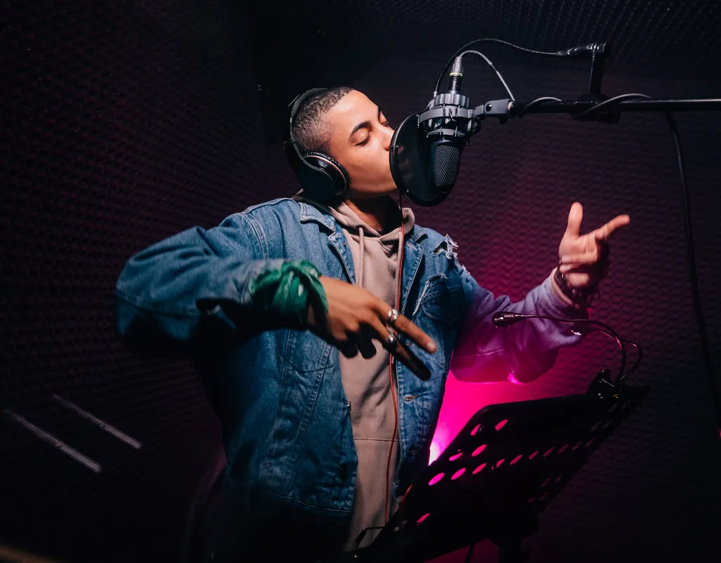 Singer performing into a studio microphone with pink and purple lighting and a music stand nearby