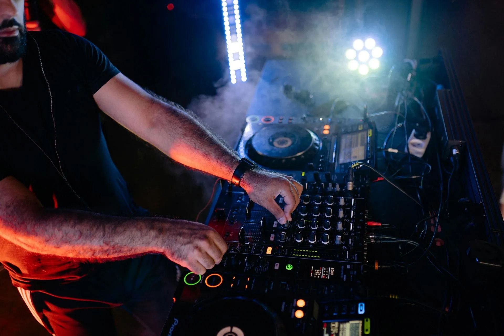 DJ mixing music on a lit turntable console in a dark nightclub, hands adjusting controls