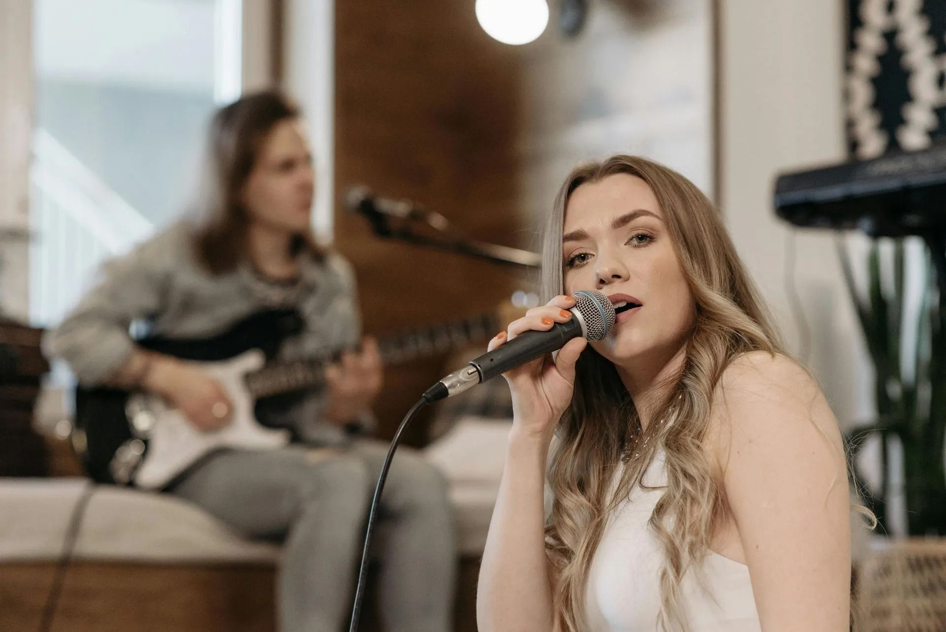 Singer performing into a microphone with a guitarist seated behind her in a studio setting