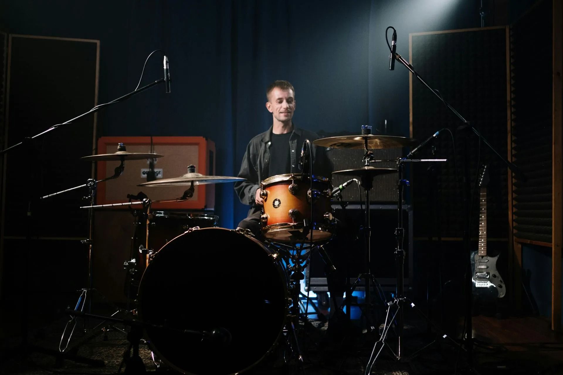 Drummer seated at a drum kit on a dark stage with microphones and amplifiers nearby
