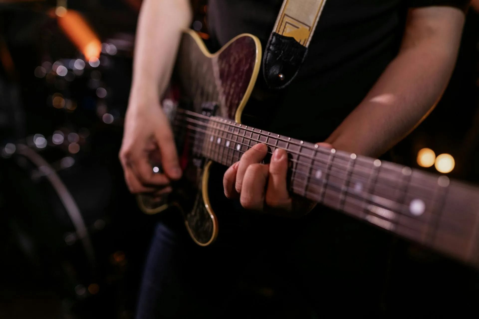 Close-up of hands playing an electric guitar on a dark stage with warm lights in the background