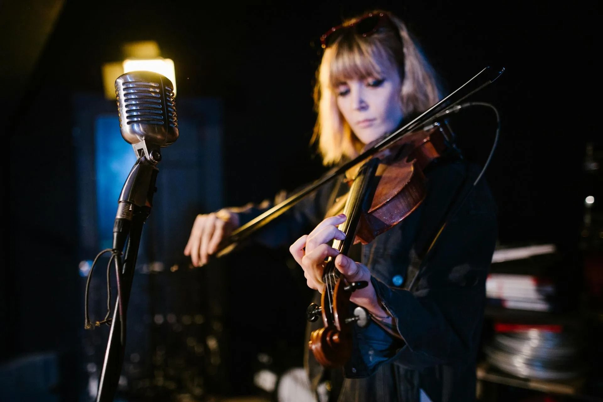 Violinist performing on stage beside a vintage microphone, lit by warm blue and amber lights