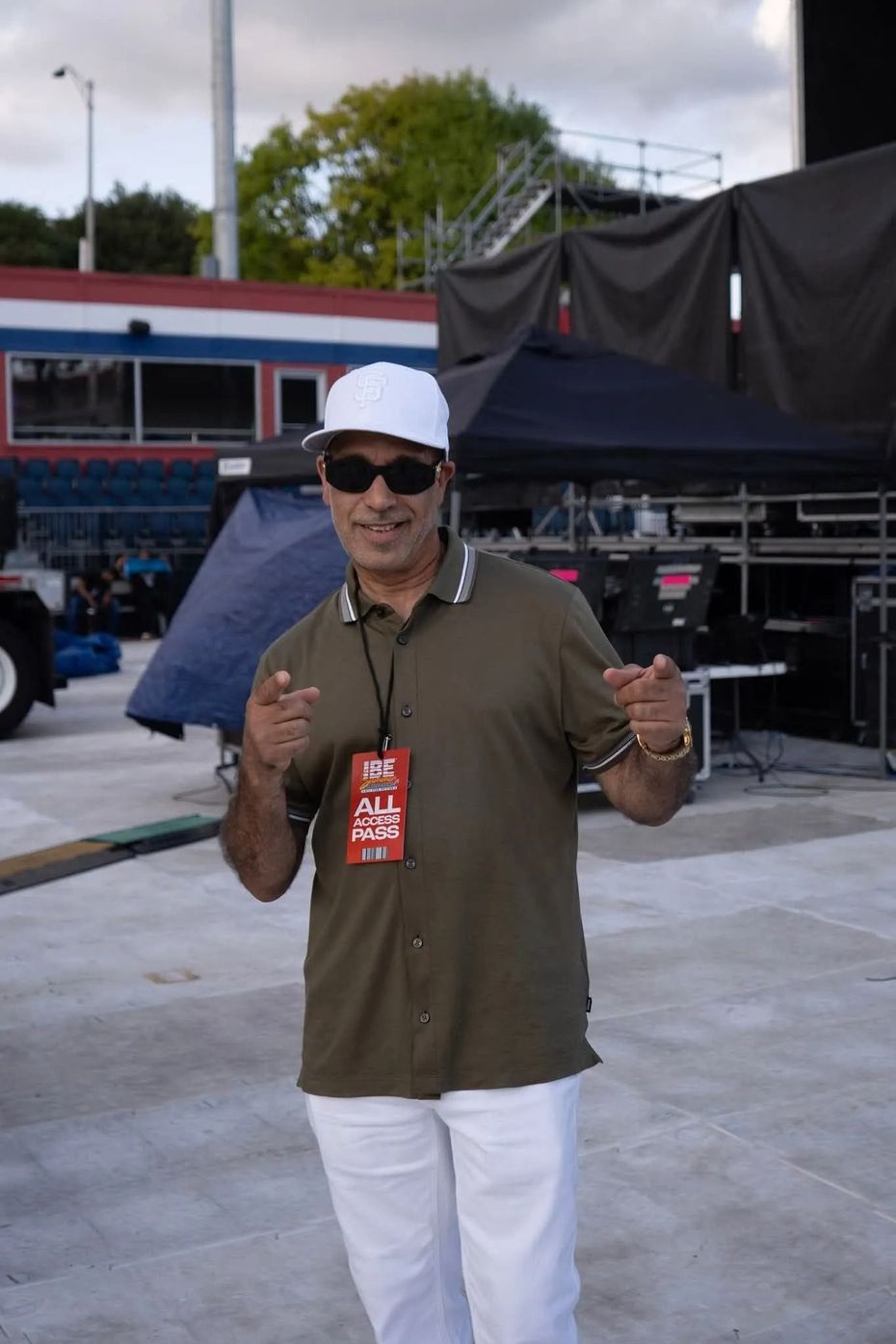Person in white cap and sunglasses standing outdoors, holding up two drinks at an event stage.