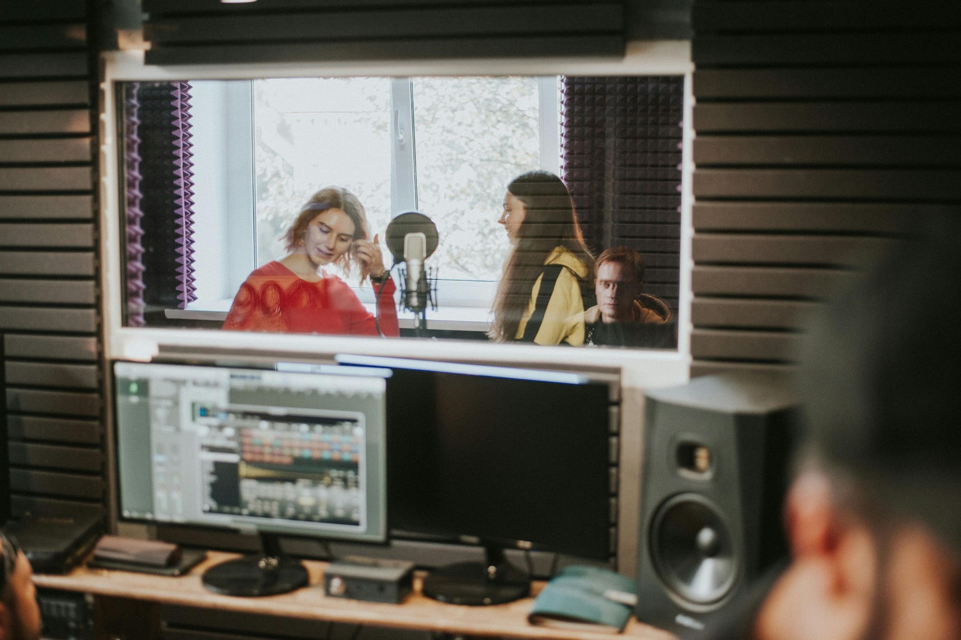 People recording a podcast in a studio, seen through a window with microphones and monitors