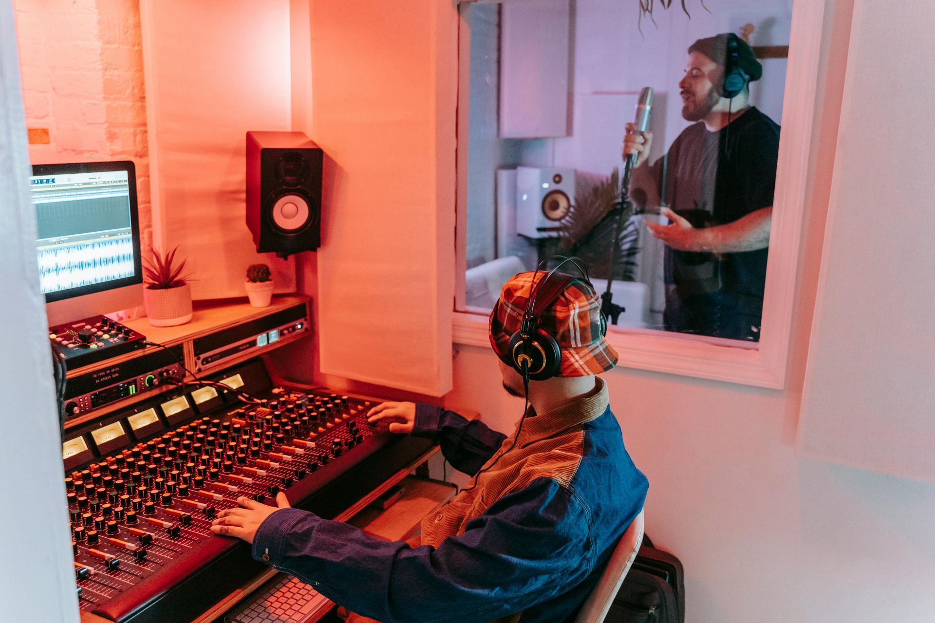 Sound engineer at a recording console with monitor and microphone in a red-lit studio, seen through a window.