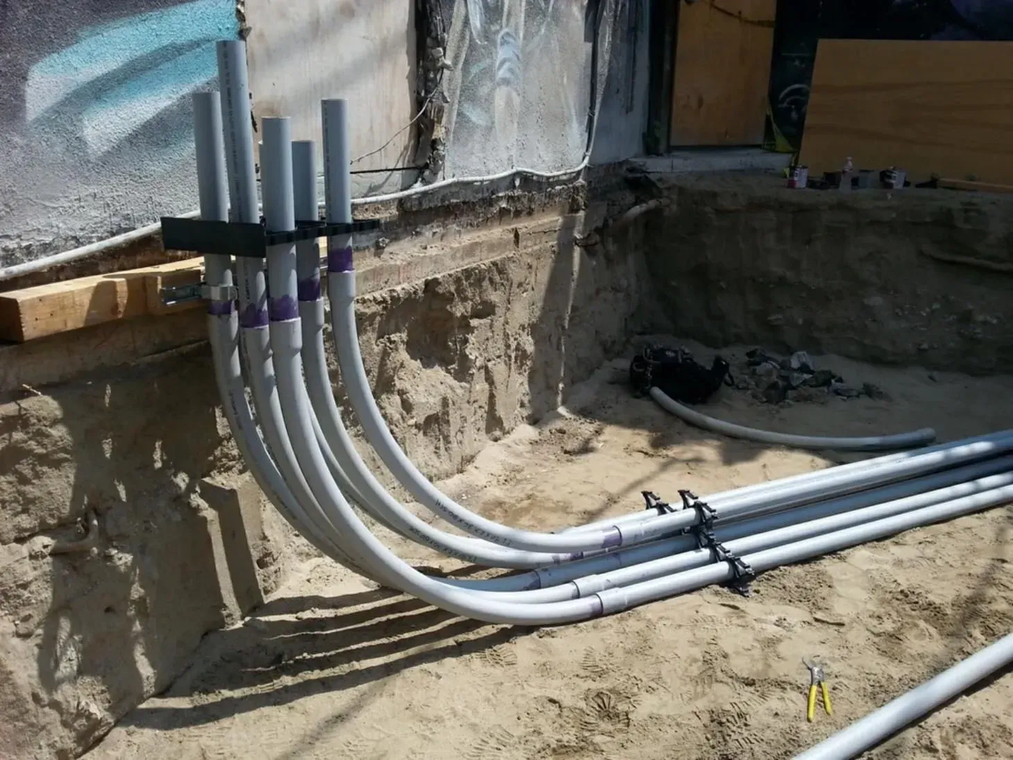 Gray electrical conduits run along a rough concrete trench at a construction site.