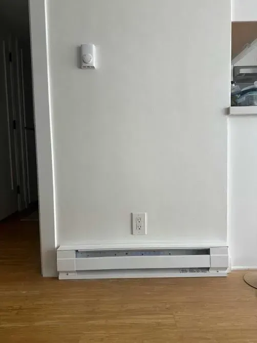 Interior wall with a baseboard heater, electrical outlet, and wooden floor near a doorway.