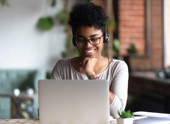 Woman wearing glasses and headphones smiles while looking at a laptop.