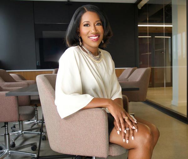 Dr. Kristin Carothers  in a beige top and skirt sits in a modern office chair, smiling.
