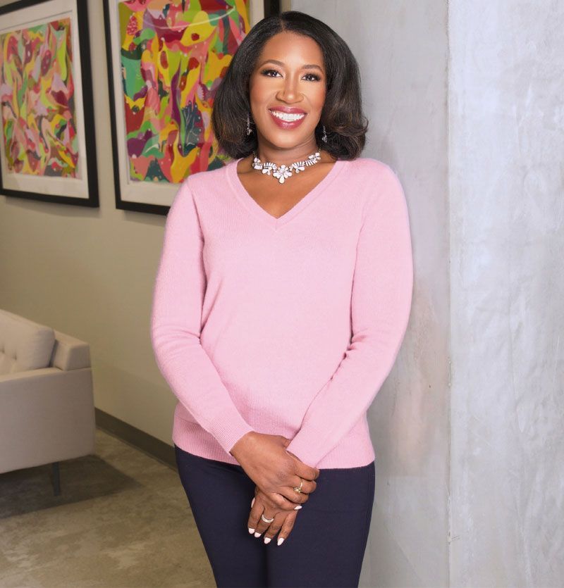 Dr. Kristin Carothers in a pink sweater and navy pants smiles, posed indoors with artwork on the wall.