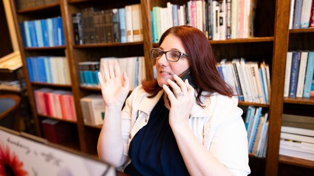 A woman is sitting in front of a bookshelf talking on a cell phone.