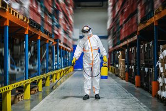 Persona in tuta protettiva che spruzza insetticida nel corridoio di un magazzino.