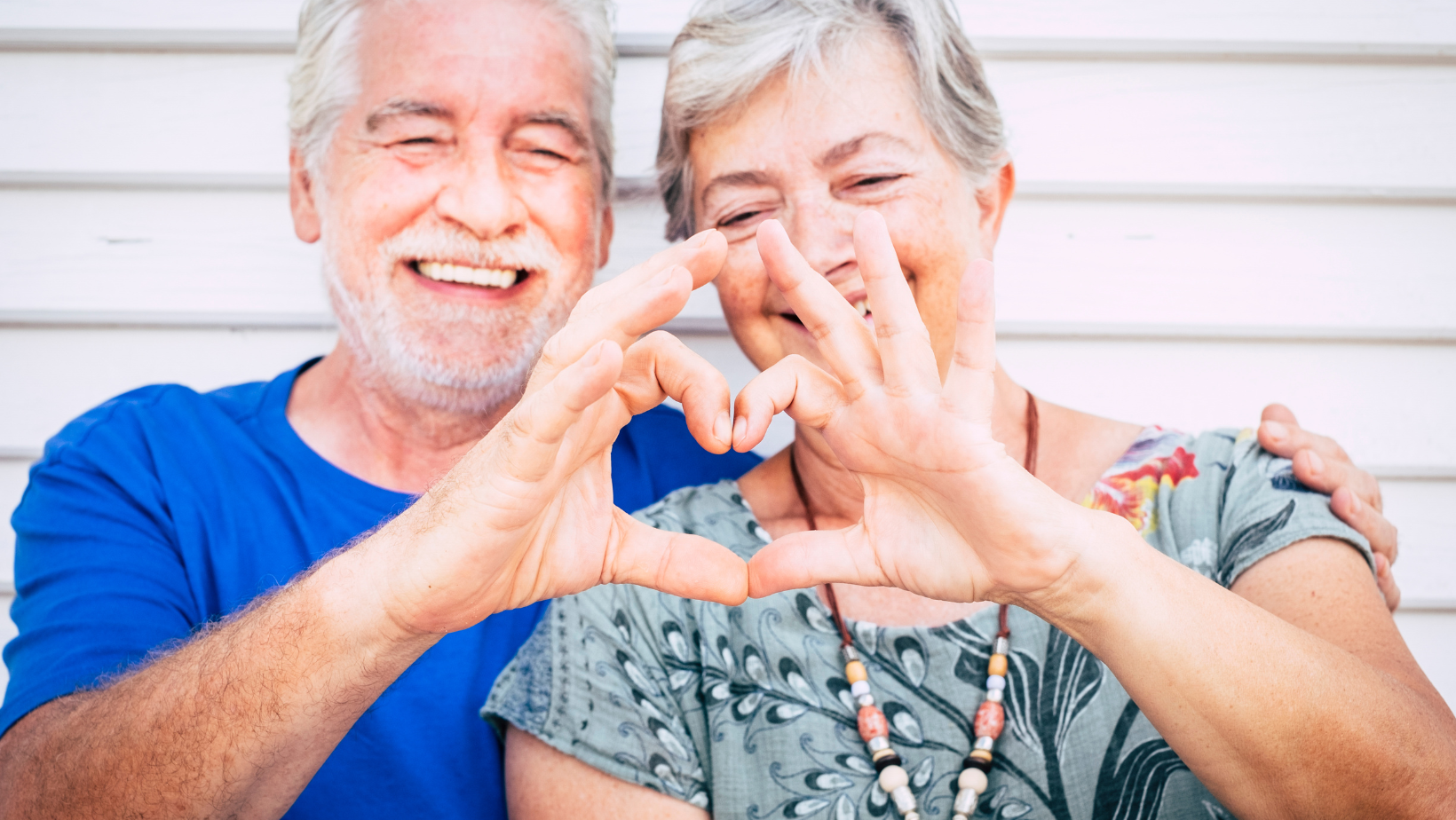 Smiling couple in blue and patterned tops forming a heart with their hands indoors