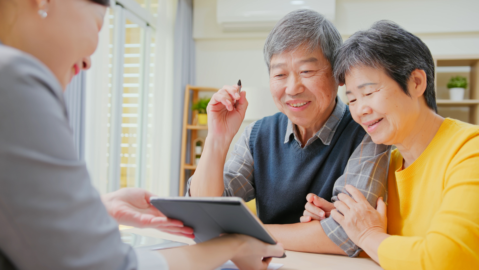 Two people smiling at a tablet while another hands them a brochure in a bright home setting