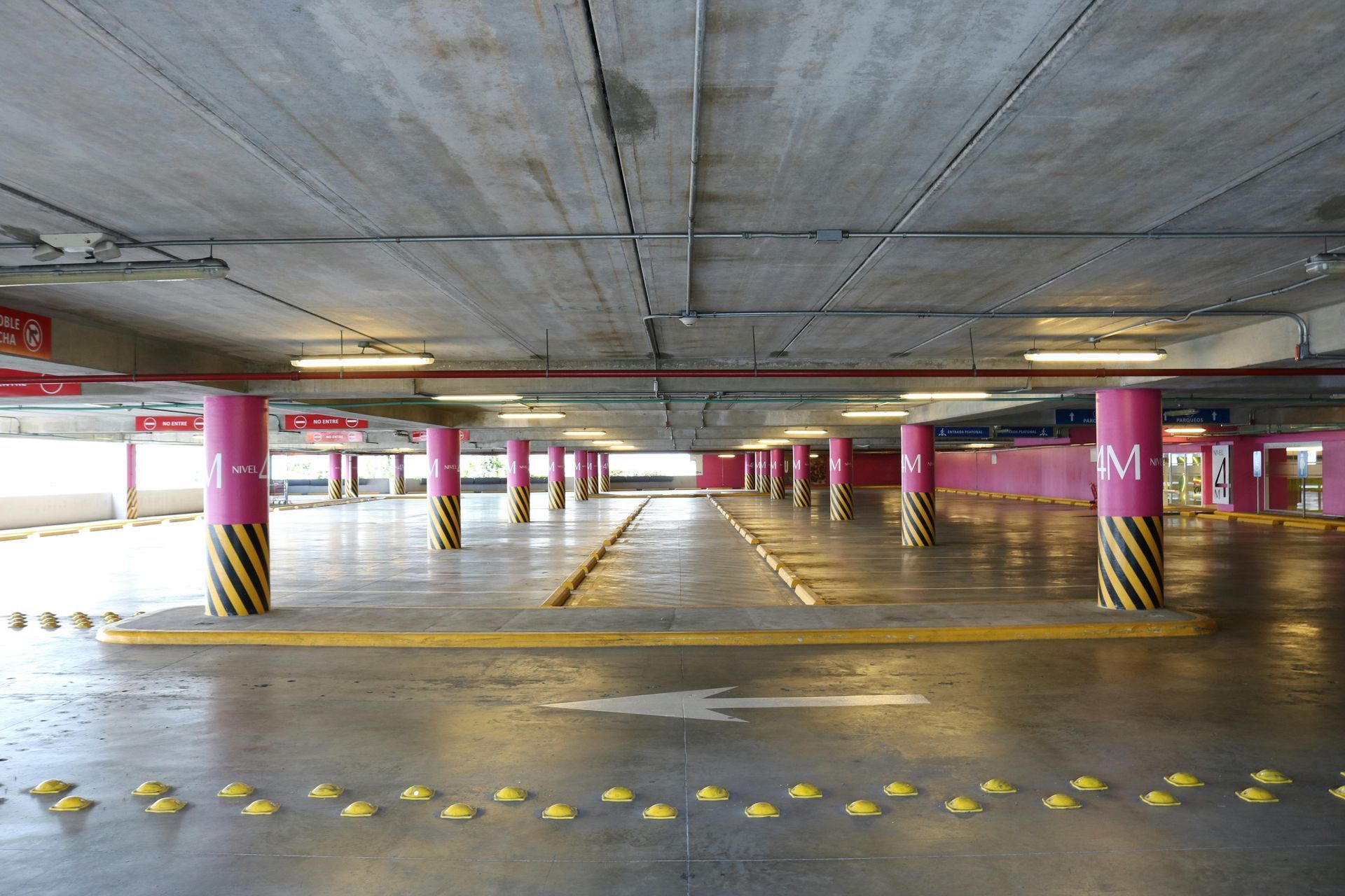 Estacionamento coberto com colunas cor-de-rosa, faixas demarcadas em amarelo e setas de direção.