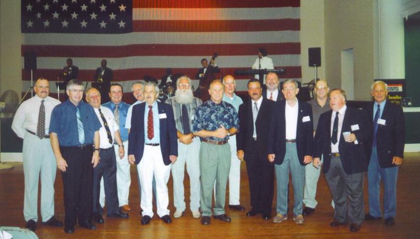 Group of men in a hall with American flag backdrop, some with name tags. A band is playing in the background.