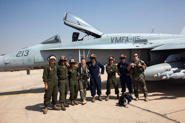 Pilots in flight suits pose in front of a gray fighter jet on a desert airfield.