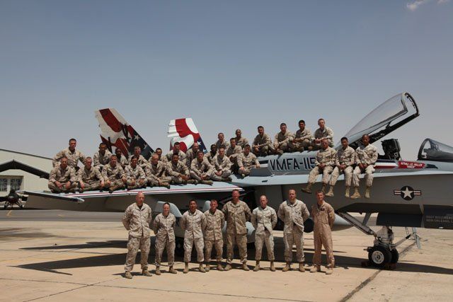 Marines in desert uniforms pose on and in front of an F/A-18 fighter jet on a sunny airfield.