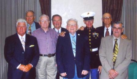 Group of nine men; some in suits, one in Marine uniform, posing indoors.