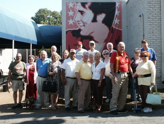 Group of people posing in front of a mural of a man; building with blue awning; sunny day.
