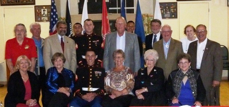 Group of people posing for a photo, likely a veterans gathering, with flags in the background.