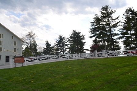 White building and fence on a grassy hill with trees under a cloudy sky.