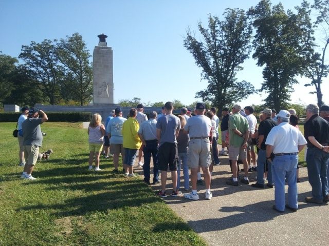 Group of people gathered outdoors, facing a tall monument. Sunny day, green grass, trees in background.
