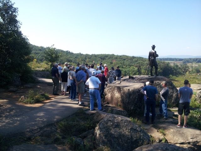 Group of people at a Gettysburg National Military Park monument, looking at statue on a rock, scenic view in background.