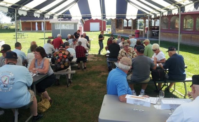 People seated at tables under a tent outdoors, with buildings and a train in the background.