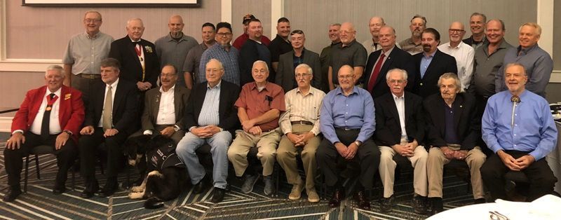 Group of men seated and standing together in a room. Many are wearing jackets and shirts.