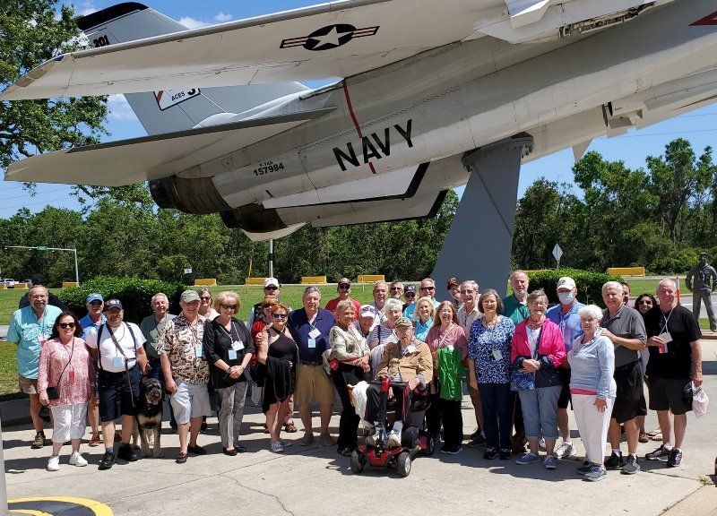 Group of people posing in front of a Navy airplane. Sunny day; trees and sky in the background.