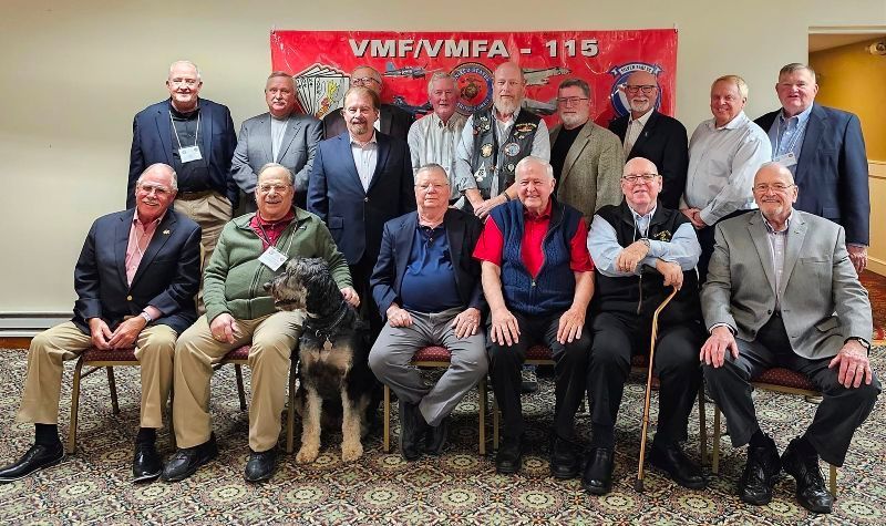 Group of men posing for a photo in front of a banner. Some are seated, and some are standing.