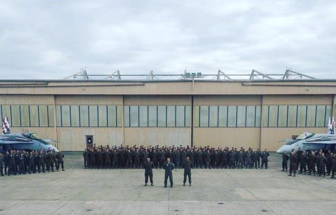 Military personnel standing in formation on an airfield, flanked by fighter jets, in front of a hangar.