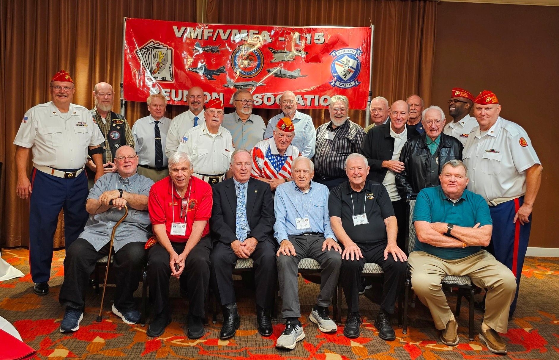 Group of men, mostly older, pose together in front of a banner. Many wear uniforms or patriotic attire.