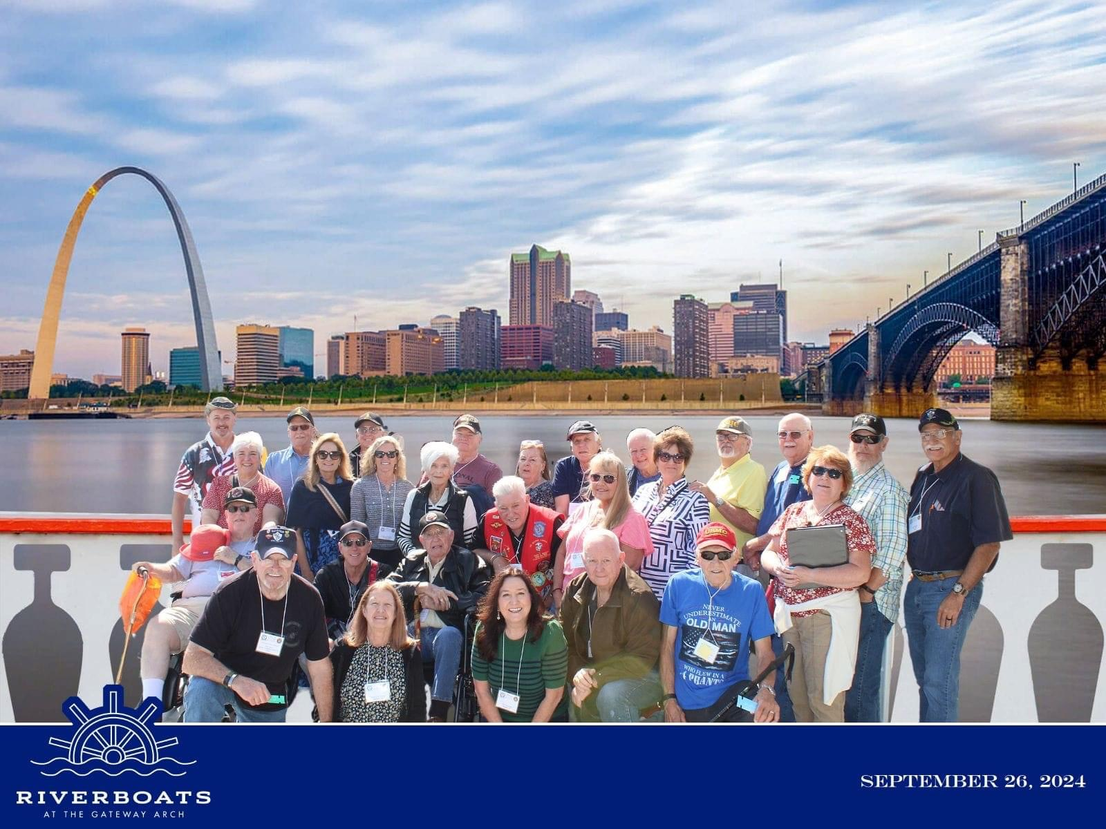 Group photo on a boat in front of the St. Louis skyline with the Gateway Arch and Eads Bridge visible.