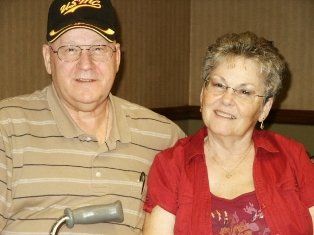 Smiling couple in tan and red shirts, glasses, and a hat. The man holds a walking cane.