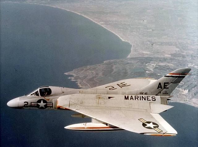 A white Marine F-4 Phantom II jet in flight over a coastal landscape.