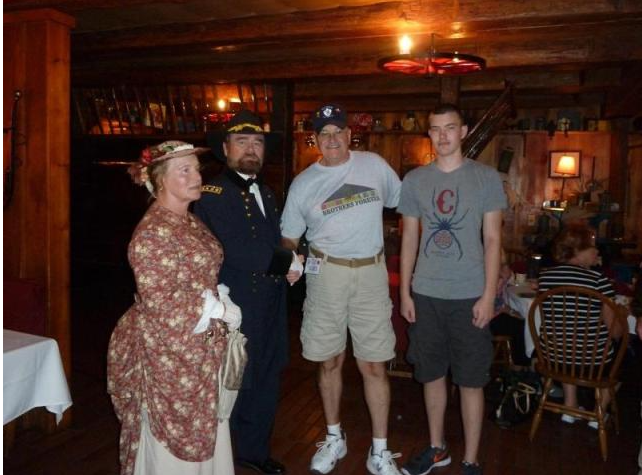 Four people pose indoors, two in period attire. Interior has wooden walls/ceiling, tables/chairs.