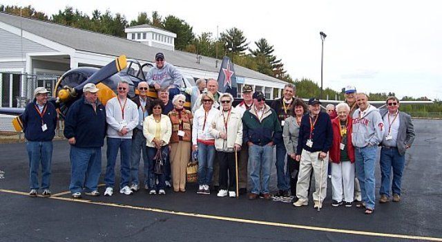 Group of people posing by a small airplane at a building with windows. They are wearing casual clothing.