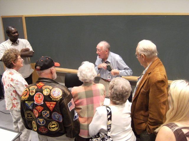 Group of people listening to a man speaking at a lectern near a chalkboard.