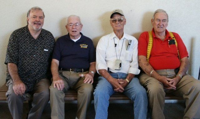 Four older men sitting on a bench. One wears a patterned shirt, the others have collared shirts, with suspenders on the far right.