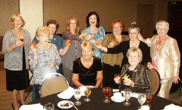 Group of women smiling, posing at a formal event, some holding small flower decorations.
