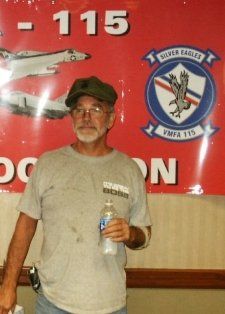 Man in a cap, holding a water bottle, stands in front of a sign with plane images and a squadron emblem.