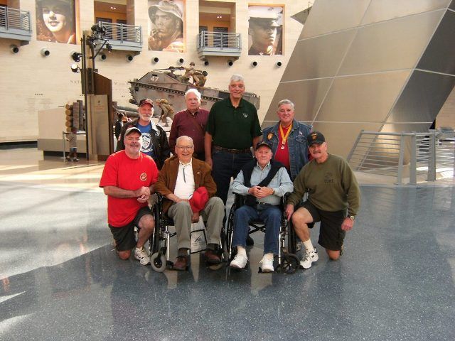 Group of veterans in a modern building. Some are in wheelchairs. Memorial in the background.