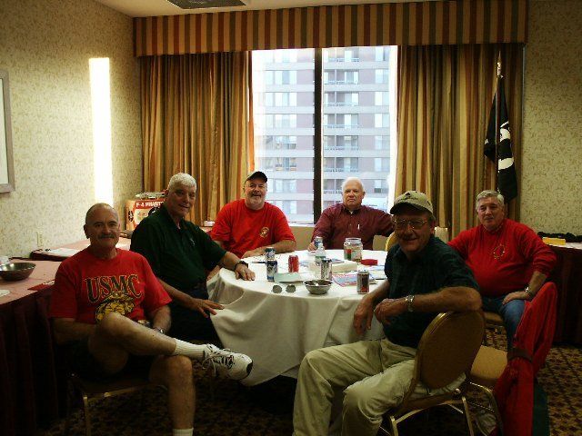 Six men seated around a table, engaged in a group activity, in a hotel room with city views.
