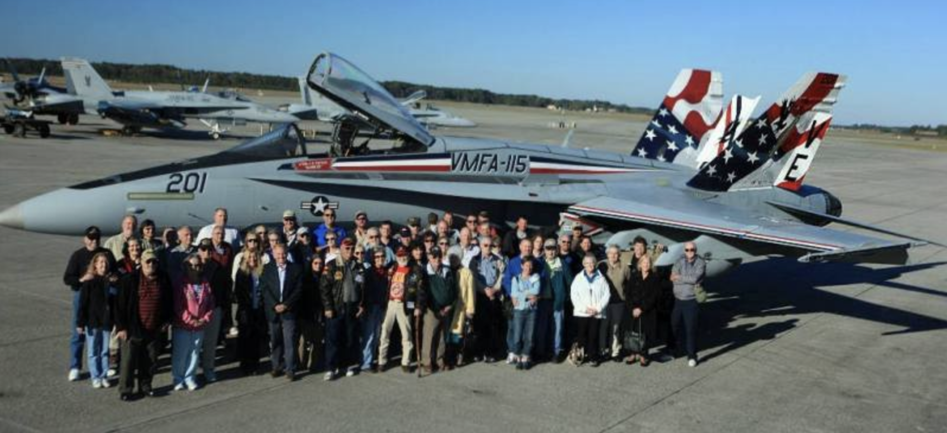Group of people posing with a fighter jet on an airfield. The jet has an American flag tail design.